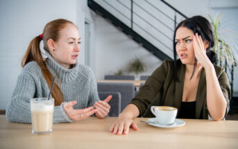 Two women sit at a table in a cozy setting, engaged in an intense conversation. The woman on the left has red hair, is wearing a gray sweater, and gestures expressively. The woman on the right has dark hair, appears frustrated, and rests her hand on her head. They have coffee drinks in front of them.