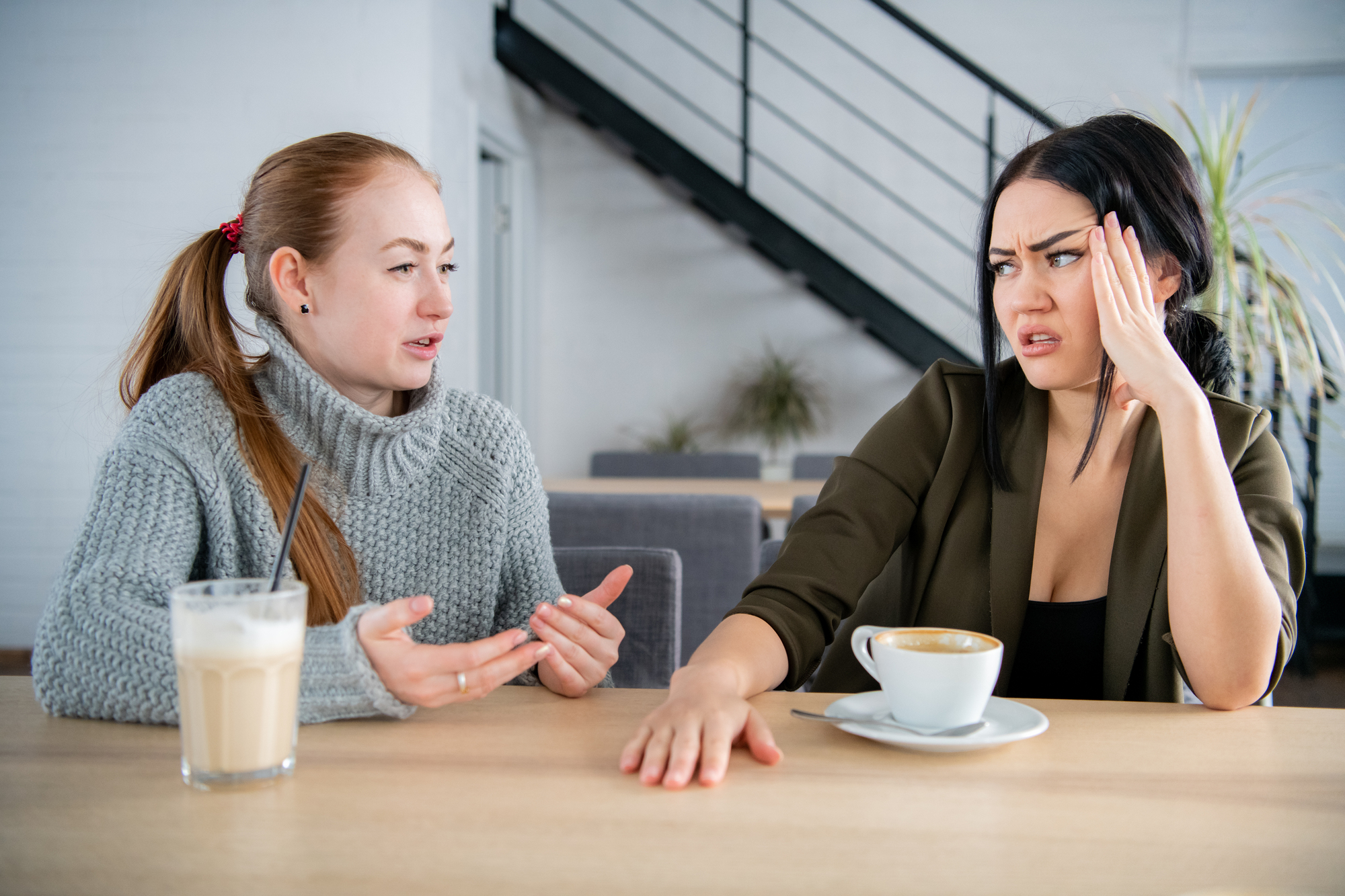 Two women sit at a table in a cozy setting, engaged in an intense conversation. The woman on the left has red hair, is wearing a gray sweater, and gestures expressively. The woman on the right has dark hair, appears frustrated, and rests her hand on her head. They have coffee drinks in front of them.