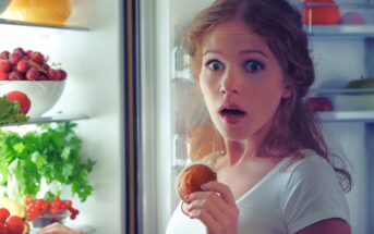 A surprised woman with curly hair and a white shirt stands by an open refrigerator, holding a bitten apple near her mouth. Her wide-eyed expression suggests shock or amazement. Inside the fridge are visible fruits, vegetables, and greens.