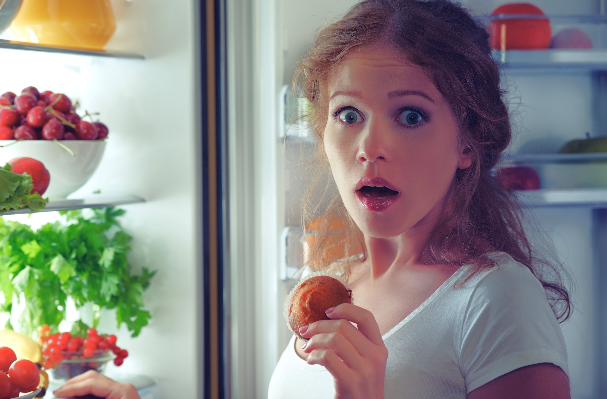 A surprised woman with curly hair and a white shirt stands by an open refrigerator, holding a bitten apple near her mouth. Her wide-eyed expression suggests shock or amazement. Inside the fridge are visible fruits, vegetables, and greens.