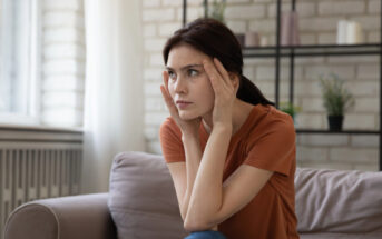 A woman sits on a couch with a concerned expression, holding her temples with both hands. She is wearing an orange shirt and has brown hair tied back. In the background, there is a brick wall, a radiator, and shelves with plants and decor items.