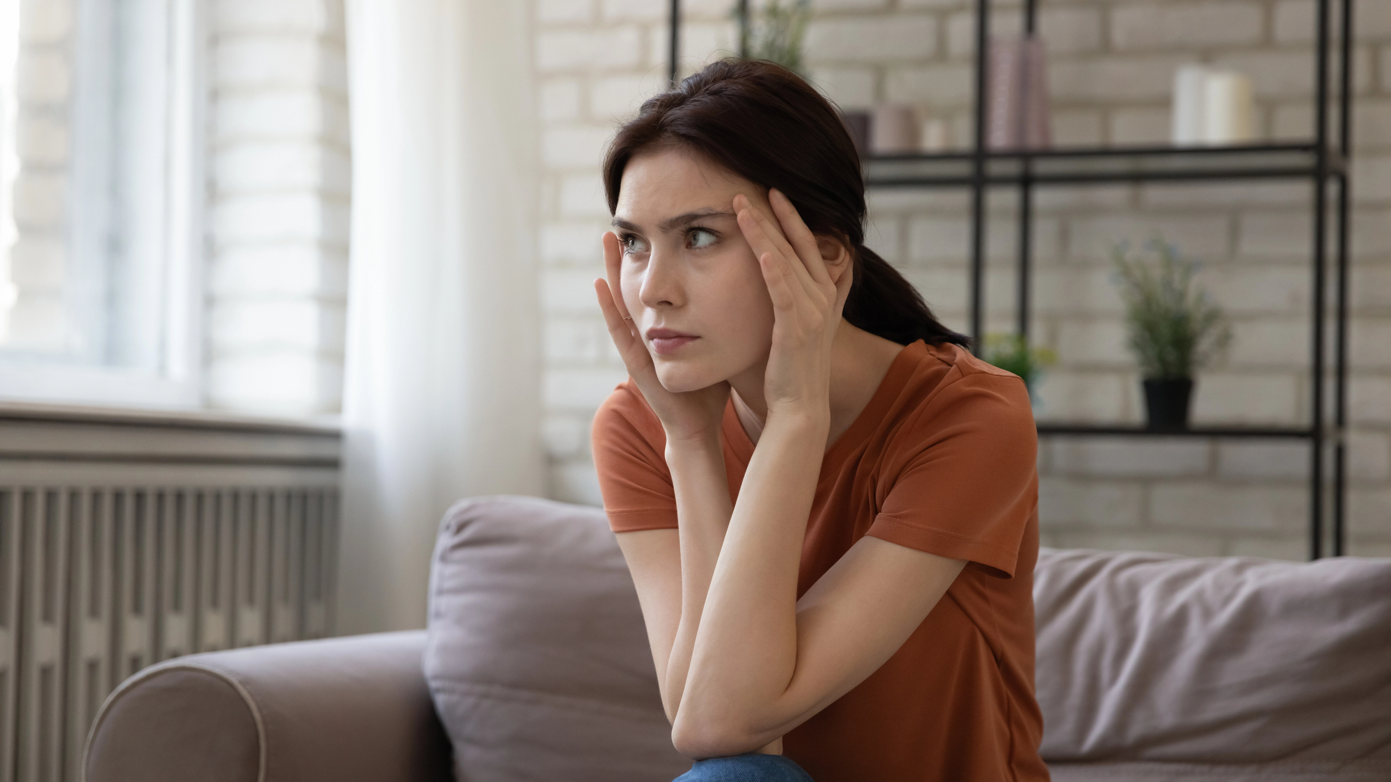 A woman sits on a couch with a concerned expression, holding her temples with both hands. She is wearing an orange shirt and has brown hair tied back. In the background, there is a brick wall, a radiator, and shelves with plants and decor items.