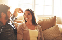 A man and woman sitting on a couch by a window, smiling at each other. The man is playfully touching the woman's hair, and they appear to be in a cheerful mood, enjoying the sunlight streaming in from the window behind them.