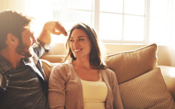 A man and woman sitting on a couch by a window, smiling at each other. The man is playfully touching the woman's hair, and they appear to be in a cheerful mood, enjoying the sunlight streaming in from the window behind them.