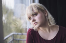 A young woman with short blonde hair and a burgundy top gazes out of a window with a contemplative expression. The background shows a blurred view of greenery and a balcony railing.