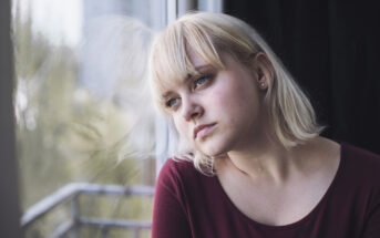 A young woman with short blonde hair and a burgundy top gazes out of a window with a contemplative expression. The background shows a blurred view of greenery and a balcony railing.