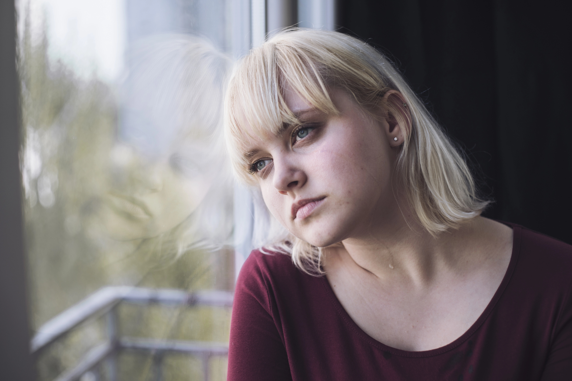 A young woman with short blonde hair and a burgundy top gazes out of a window with a contemplative expression. The background shows a blurred view of greenery and a balcony railing.