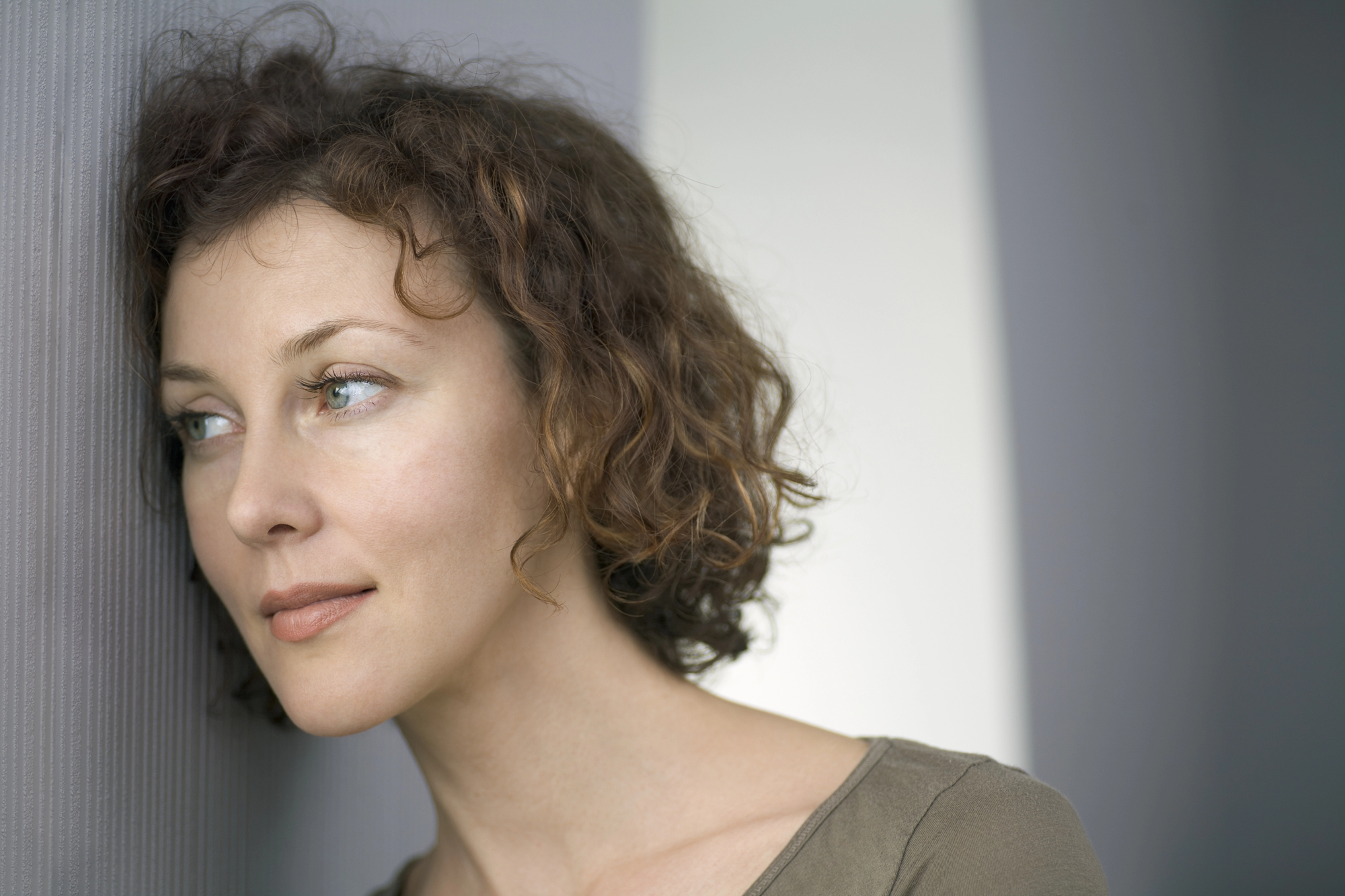 A woman with short curly brown hair leans her head against a textured wall, looking off to the side with a thoughtful expression. She is wearing a simple olive green top, and the background consists of neutral gray and white tones.