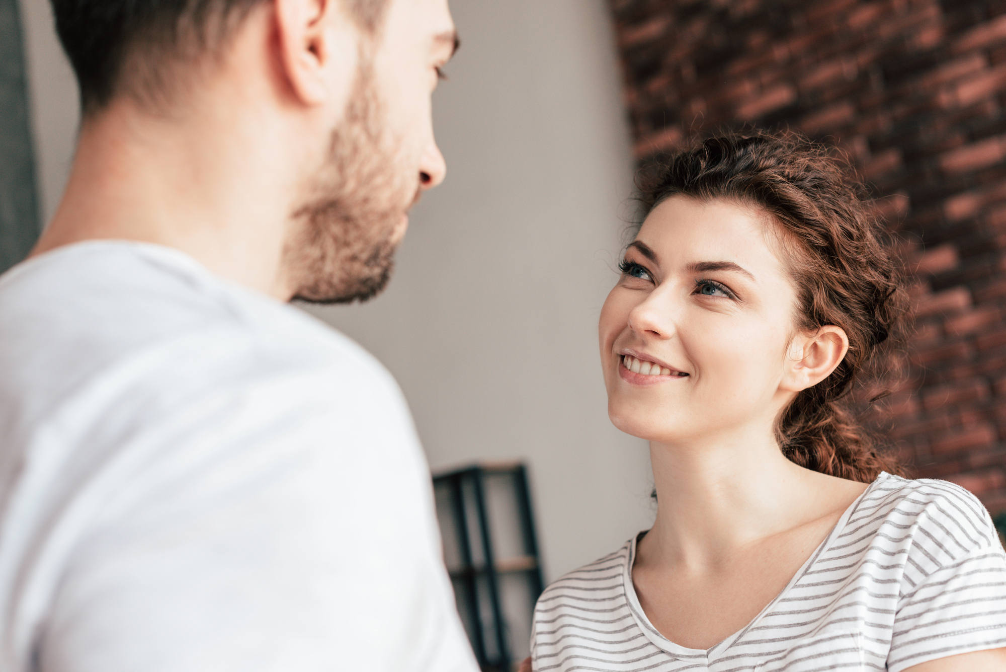 A woman with curly brown hair and a striped shirt smiles while looking up at a man with a short beard. They are indoors, standing close to each other, with a brick wall partially visible in the background.