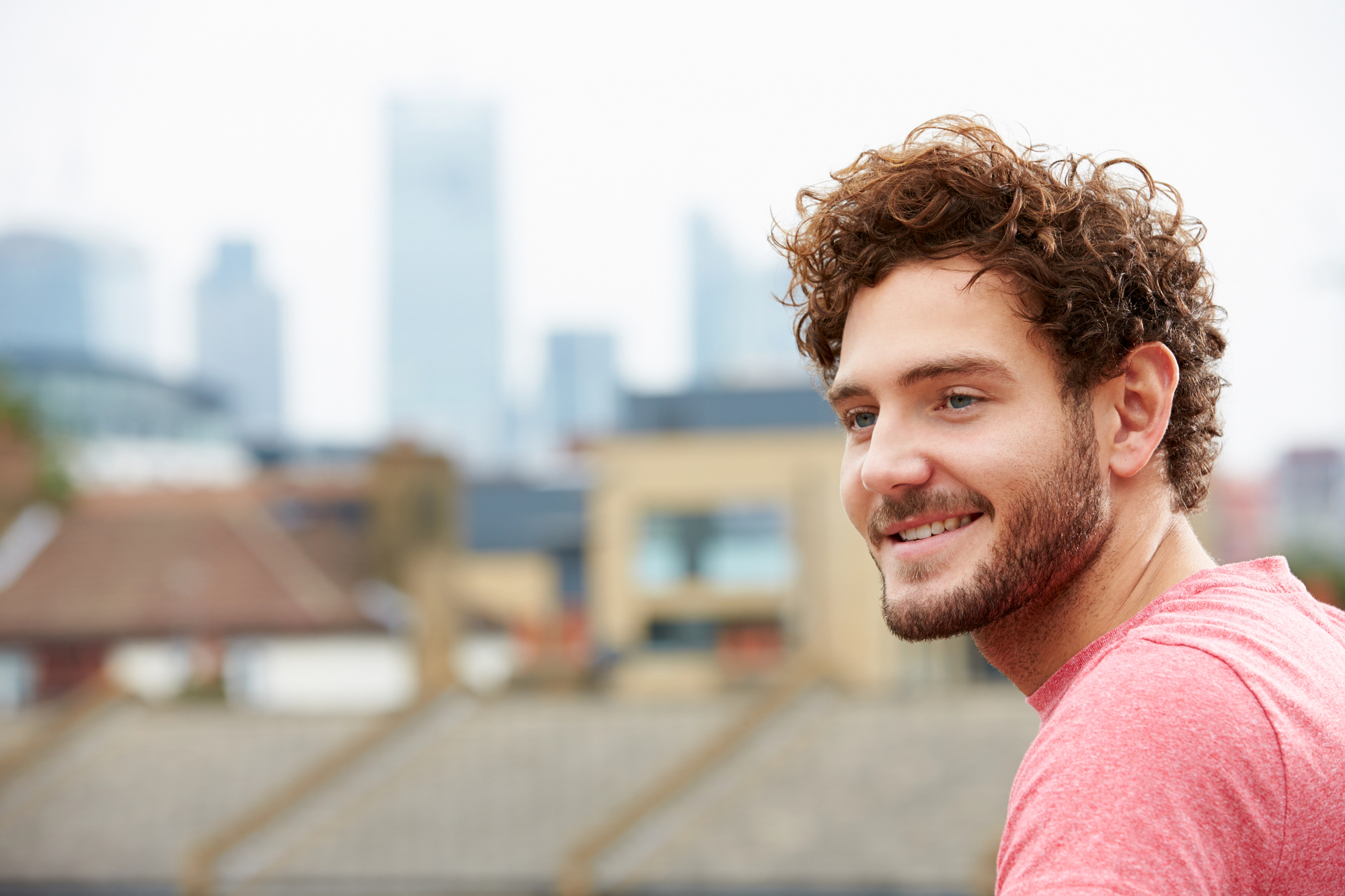 A young man with curly brown hair and a beard smiles while gazing off to the side in an urban outdoor setting with building rooftops and a city skyline blurred in the background. He is wearing a red shirt.