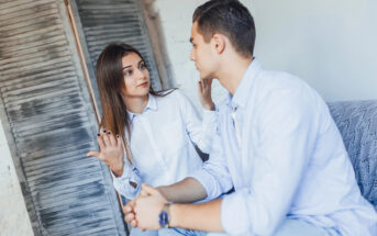 A woman and a man sit on a couch having a serious conversation. The woman gestures with her hand while looking at the man, who is listening attentively. Both are dressed in casual white shirts. A wooden shutter door is in the background.