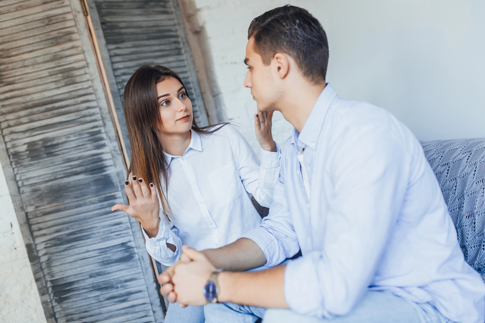 A woman and a man sit on a couch having a serious conversation. The woman gestures with her hand while looking at the man, who is listening attentively. Both are dressed in casual white shirts. A wooden shutter door is in the background.