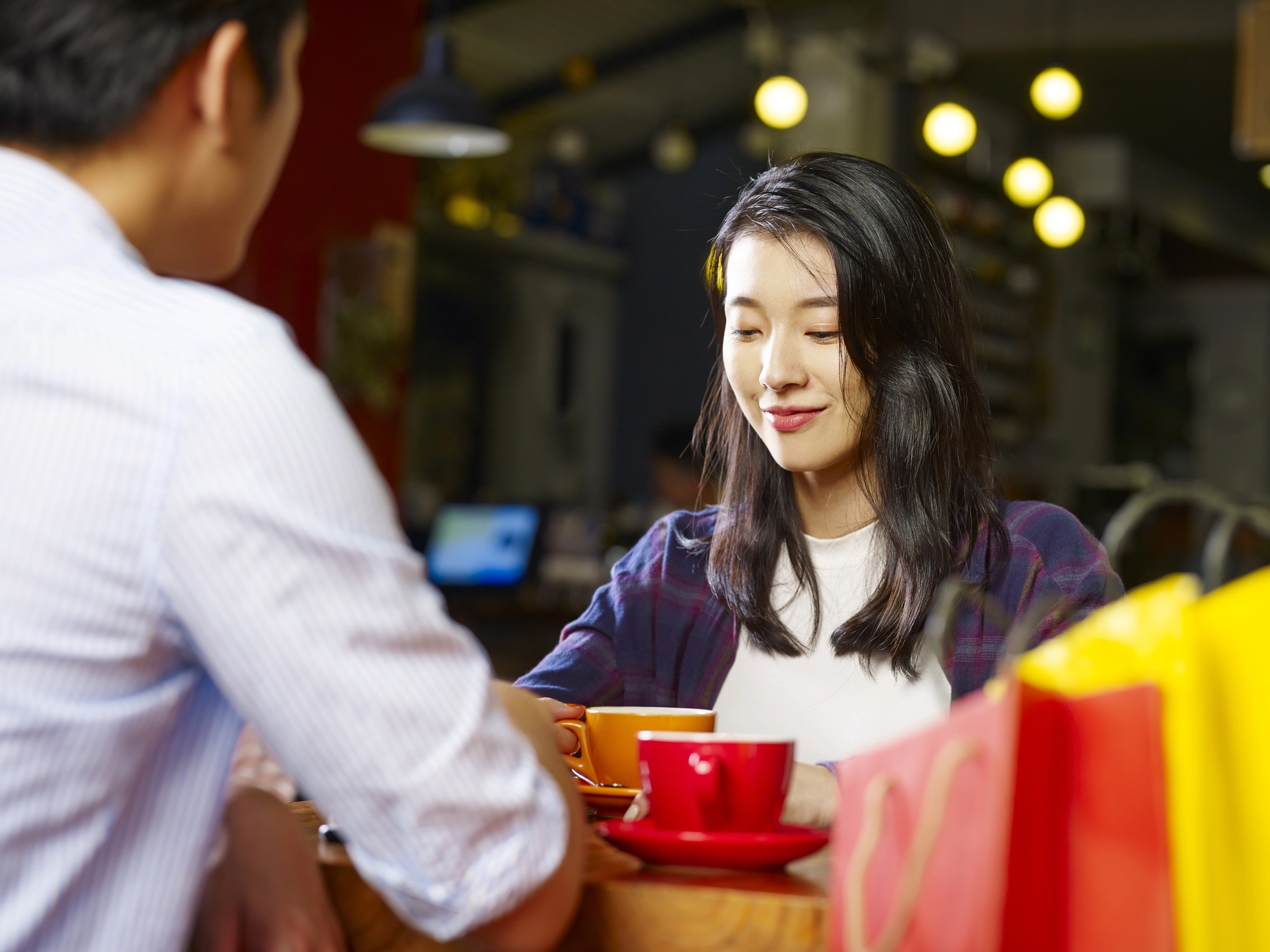 Two people are sitting indoors at a table in a cafe, enjoying coffee together. The woman on the right is smiling down at her cup, while the man on the left is partially visible, holding his cup. The setting is cozy, with warm lighting and shopping bags beside them.