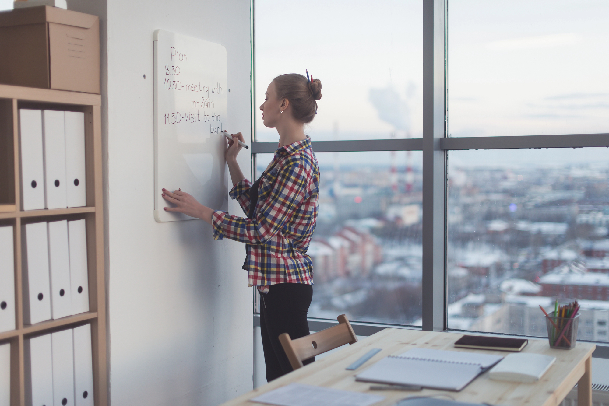 A person wearing a plaid jacket is standing and writing on a whiteboard in an office. The office has large windows showcasing a cityscape. Near a table with notebooks, a tablet, and files, there is a shelf with white binders on the left side.