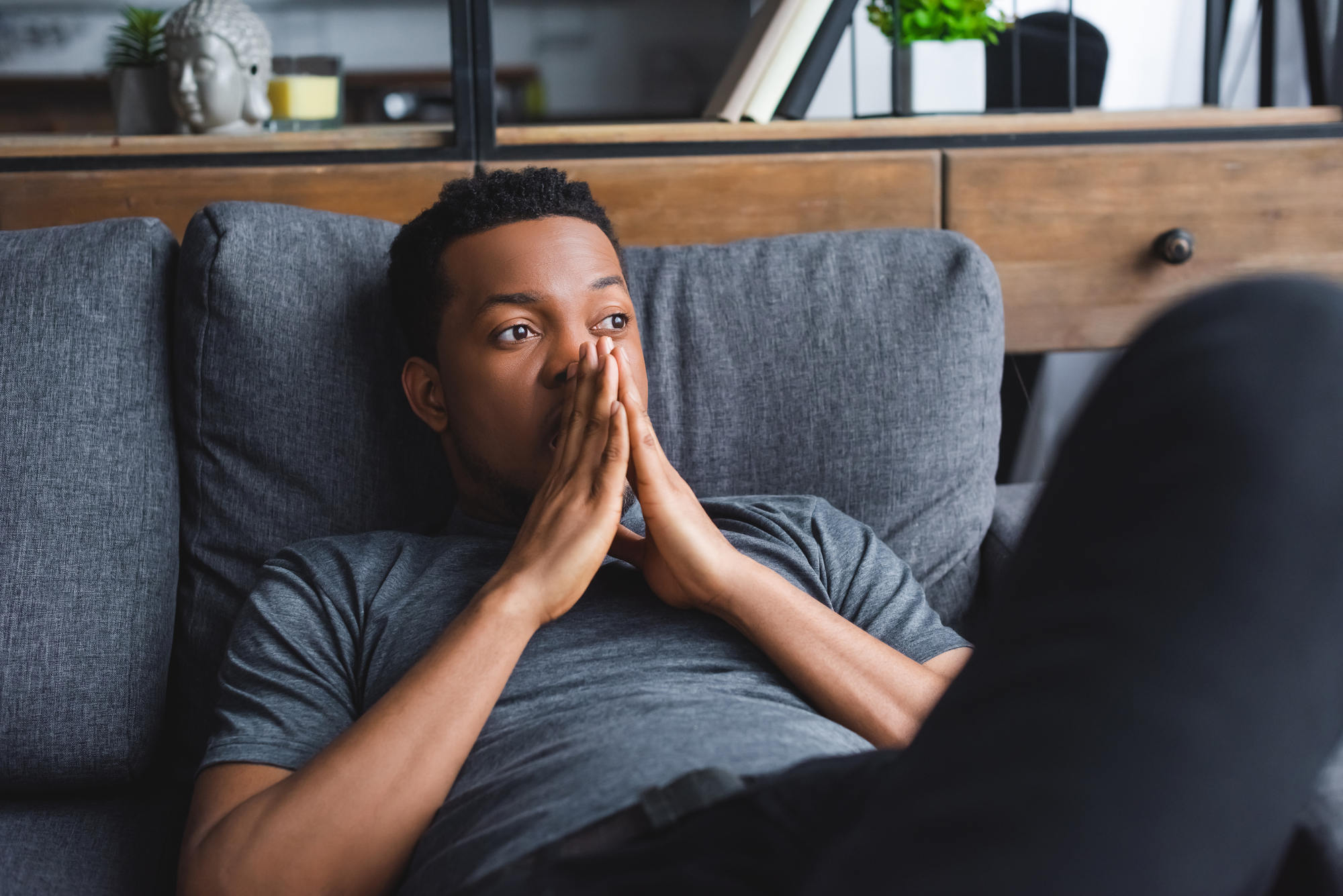 A man with short, curly hair wearing a gray t-shirt is lying on a gray couch, looking thoughtful with his hands clasped in front of his face. He appears to be deep in thought. The background features wooden furniture and a decorative item on a shelf.