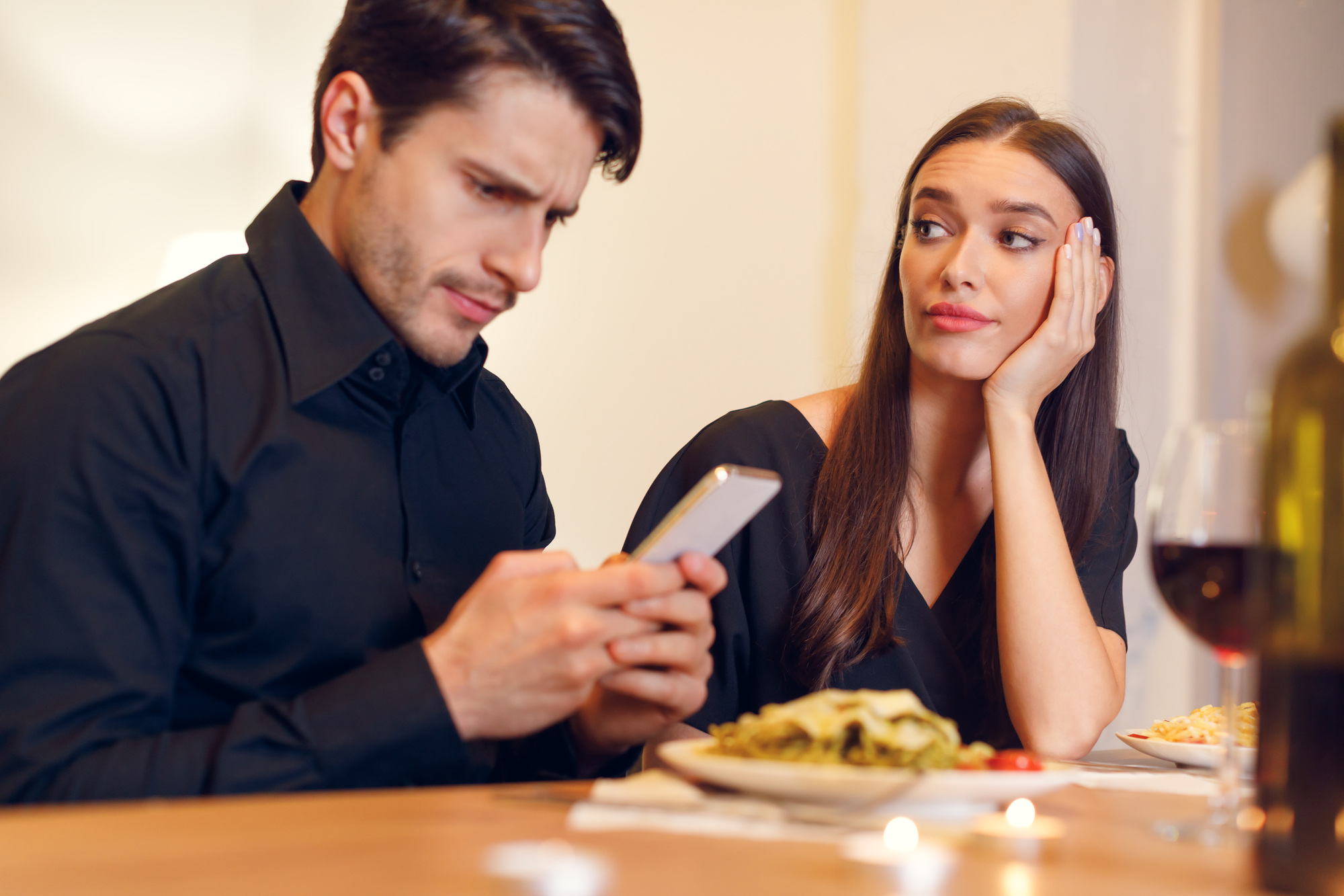 A man and woman sit at a table in a restaurant. The man is engrossed in his phone, while the woman, with her head resting on her hand, looks at him with a mix of frustration and disappointment. Plates of food and a bottle of wine are on the table.