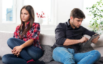 A woman and a man sit on a couch, both looking displeased. The woman, wearing a red plaid shirt and jeans, has her arms folded and looks away. The man, wearing a black shirt and jeans, looks focused on a tablet he's holding. The setting appears to be a living room.