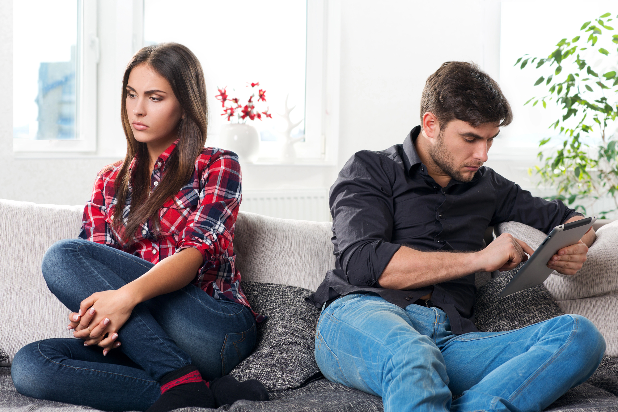 A woman and a man sit on a couch, both looking displeased. The woman, wearing a red plaid shirt and jeans, has her arms folded and looks away. The man, wearing a black shirt and jeans, looks focused on a tablet he's holding. The setting appears to be a living room.