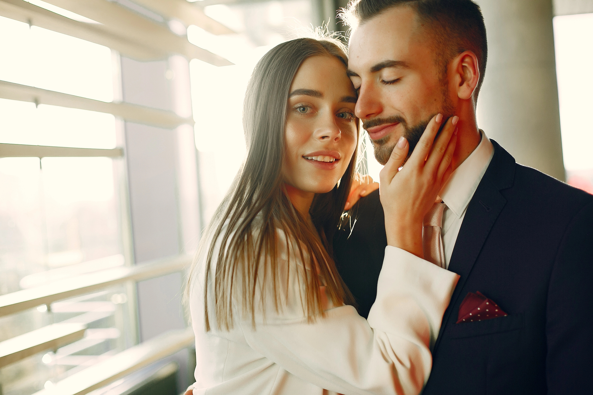 A woman with long brown hair embraces a bearded man dressed in a suit and tie. She looks at the camera while gently touching his face. The man has his eyes closed, smiling contently. They are in a brightly lit indoor setting with large windows in the background.