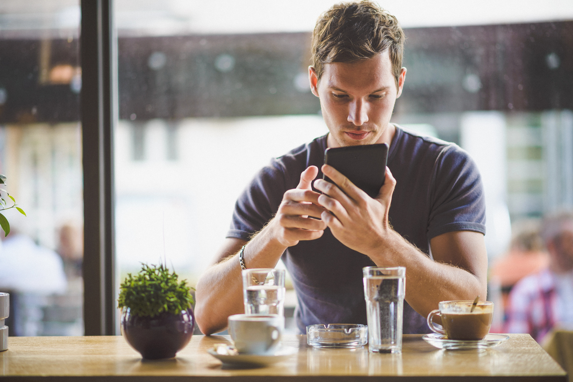 A man sits at a café table, engrossed in his smartphone. The table holds a cup of coffee, a glass of water, an ashtray, a small potted plant, and another glass. Sunlight fills the room through large windows in the background.