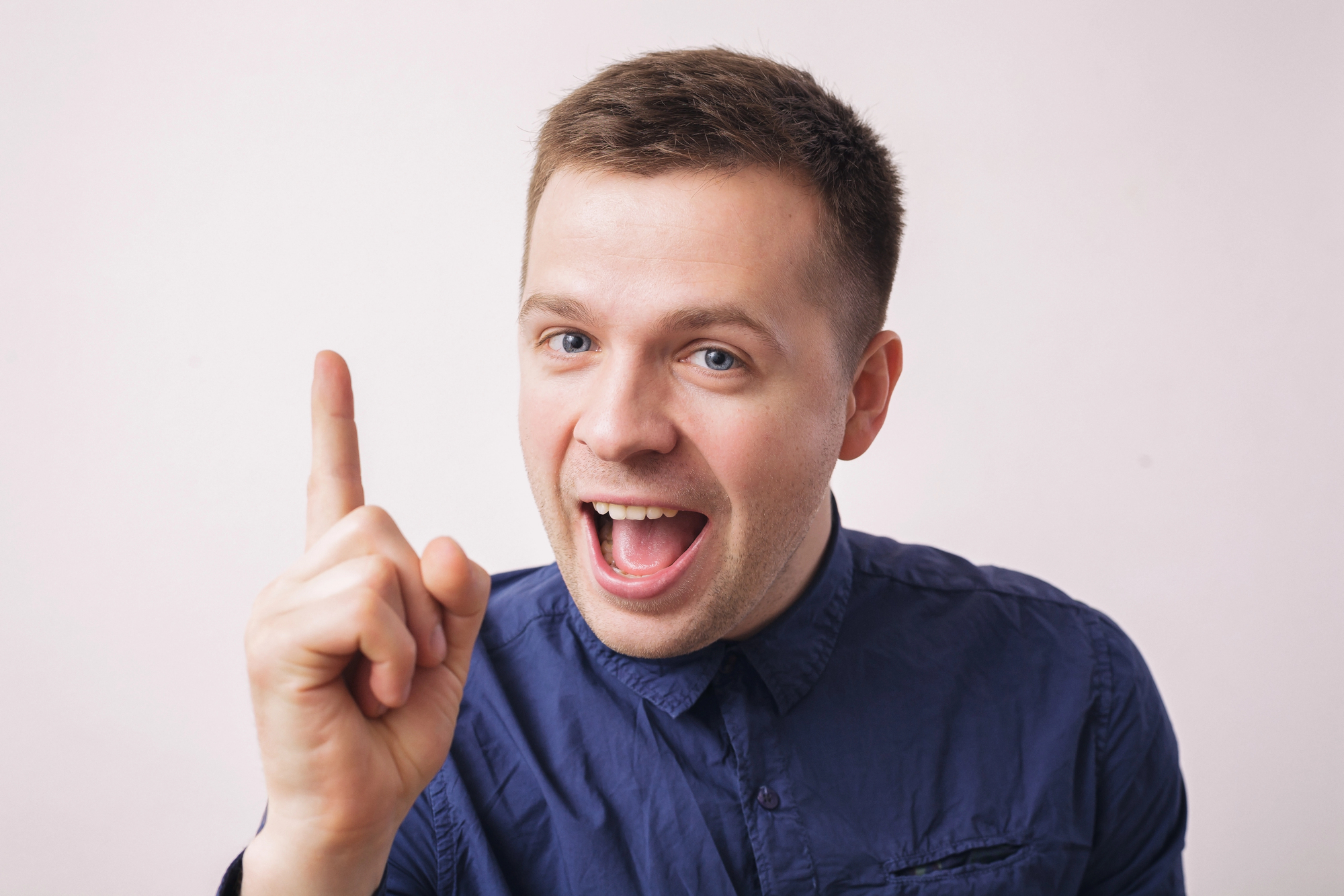 A man with short brown hair is smiling widely and pointing upwards with his right index finger. He is wearing a blue button-up shirt and standing against a plain white background.