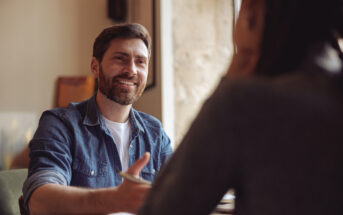 A bearded man in a denim shirt sits at a table, smiling and gesturing with his hand, while speaking to another person whose back is to the camera. They appear to be having a friendly conversation in a cozy, well-lit setting.