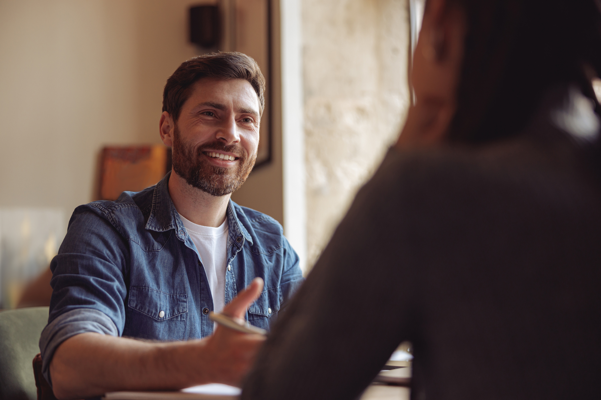 A bearded man in a denim shirt sits at a table, smiling and gesturing with his hand, while speaking to another person whose back is to the camera. They appear to be having a friendly conversation in a cozy, well-lit setting.