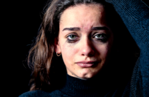 A woman with long, brown hair and green eyes stands against a dark background. She appears distressed, with smudged mascara under her eyes, indicating she has been crying. She wears a dark turtleneck sweater and has one hand raised to her head.