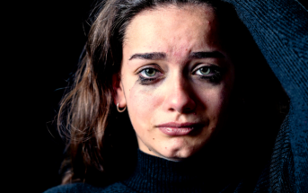 A woman with long, brown hair and green eyes stands against a dark background. She appears distressed, with smudged mascara under her eyes, indicating she has been crying. She wears a dark turtleneck sweater and has one hand raised to her head.