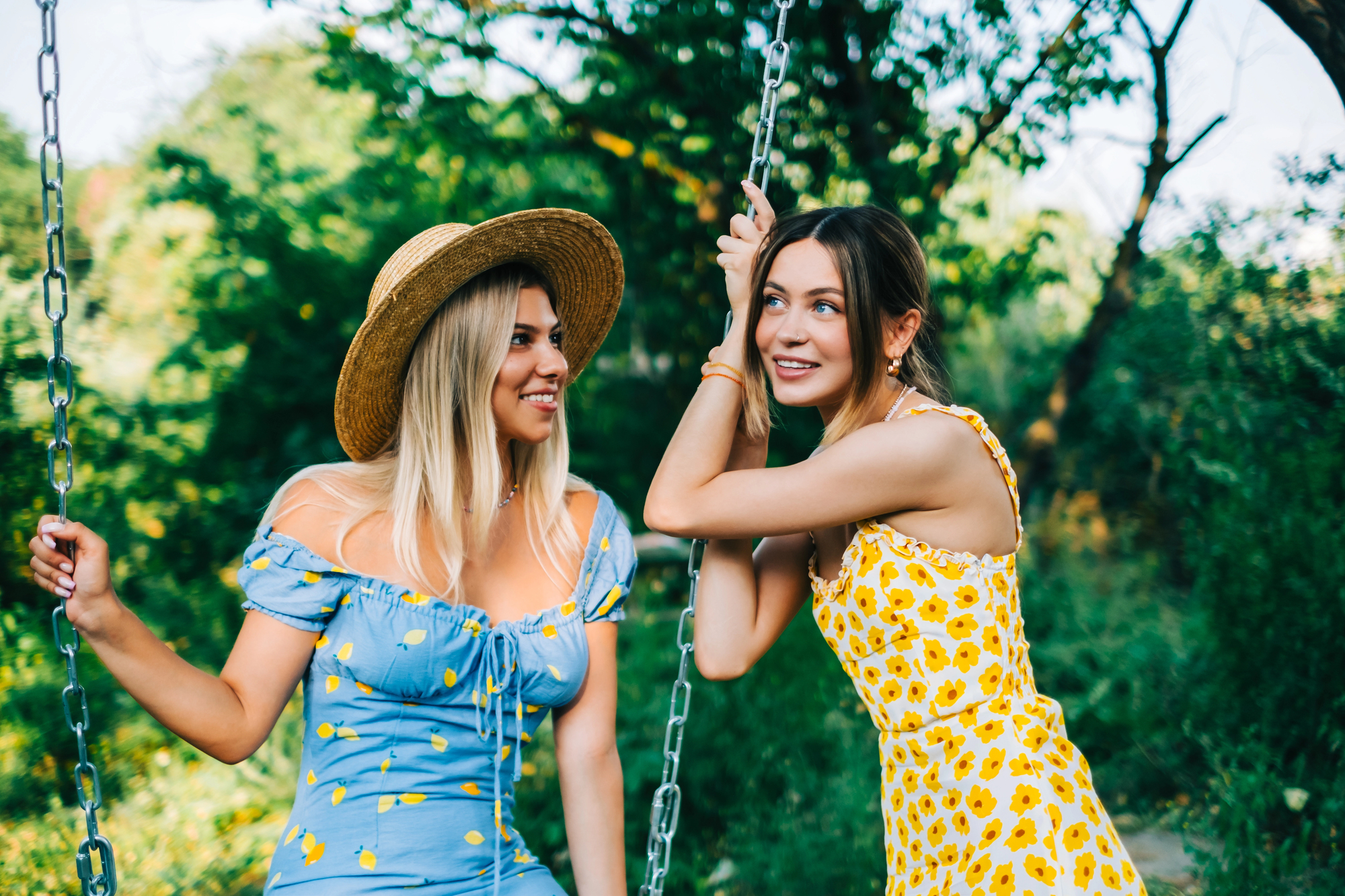 Two women are enjoying a sunny day on a swing set in a green, leafy park. One woman, wearing a blue dress and straw hat, smiles as she sits on the swing, while the other, in a yellow dress, leans against the swing chain, looking cheerful.