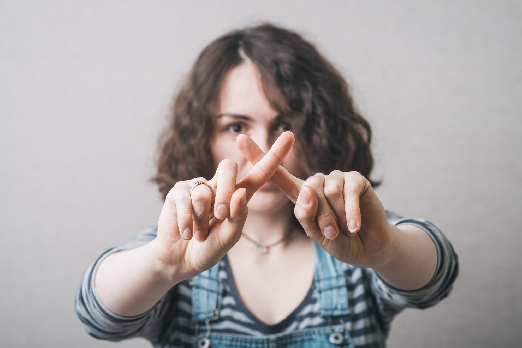 A person with curly hair is making an "X" symbol with their fingers in front of their face. They are wearing a striped shirt and denim overalls. The background is neutral.