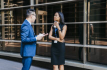 A man in a blue suit and glasses is talking to a woman in a black dress who is holding a coffee cup. They are standing outside a modern building with large windows, and both wear ID badges. The woman crosses her arms and listens attentively.