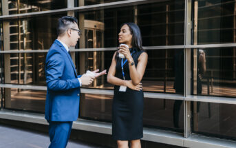 A man in a blue suit and glasses is talking to a woman in a black dress who is holding a coffee cup. They are standing outside a modern building with large windows, and both wear ID badges. The woman crosses her arms and listens attentively.