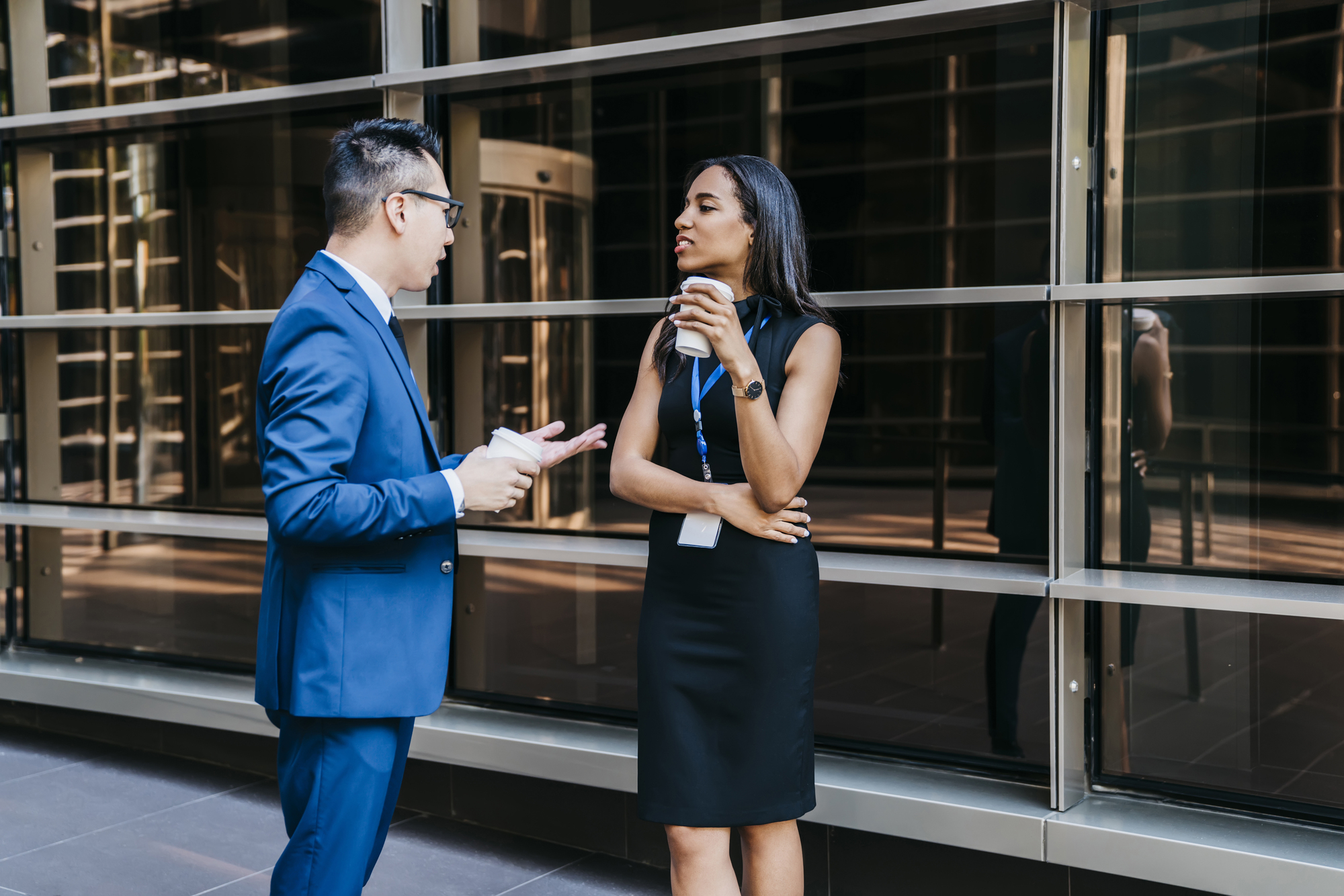 A man in a blue suit and glasses is talking to a woman in a black dress who is holding a coffee cup. They are standing outside a modern building with large windows, and both wear ID badges. The woman crosses her arms and listens attentively.