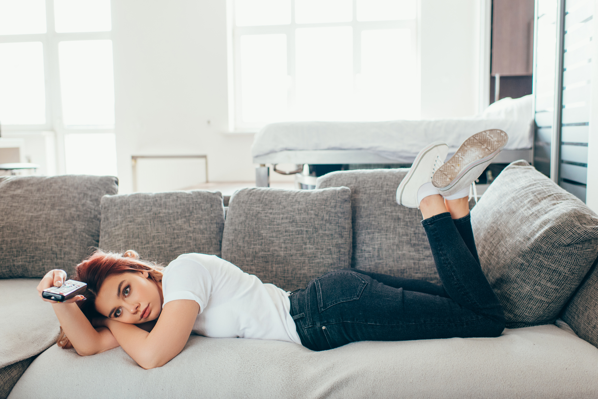 A woman with red hair is lying on a gray sofa, holding a remote control. She is wearing a white t-shirt, black jeans, and white sneakers. Her legs are propped up on the armrest. The room is bright with large windows in the background.