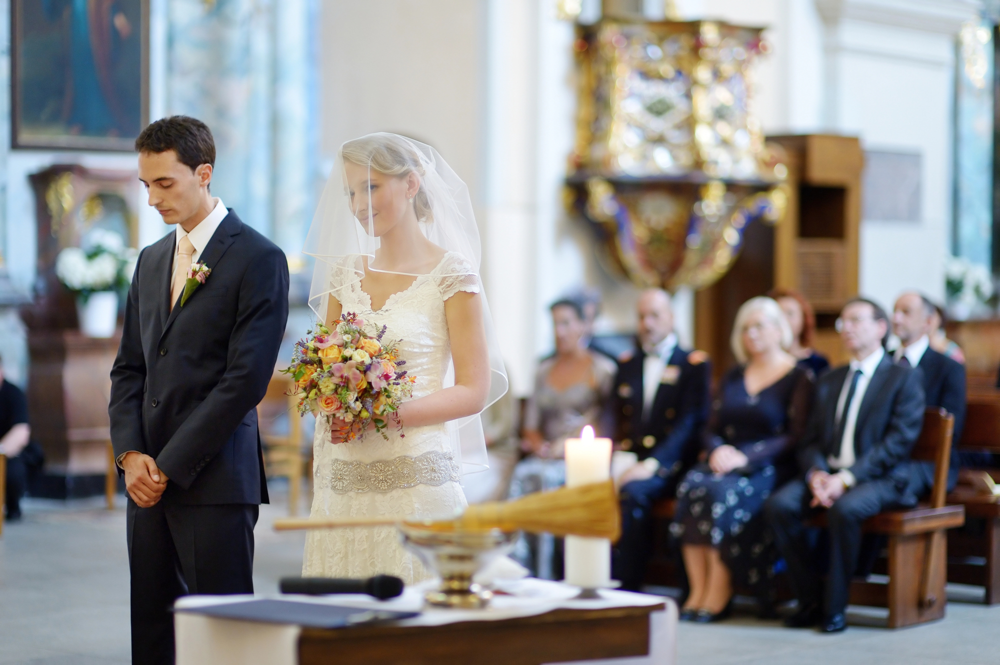 A bride and groom stand at the altar during their wedding ceremony. The bride, in a white dress and veil, holds a bouquet of flowers, while the groom, in a dark suit, looks down. In the background, guests are seated attentively in a decorated church.