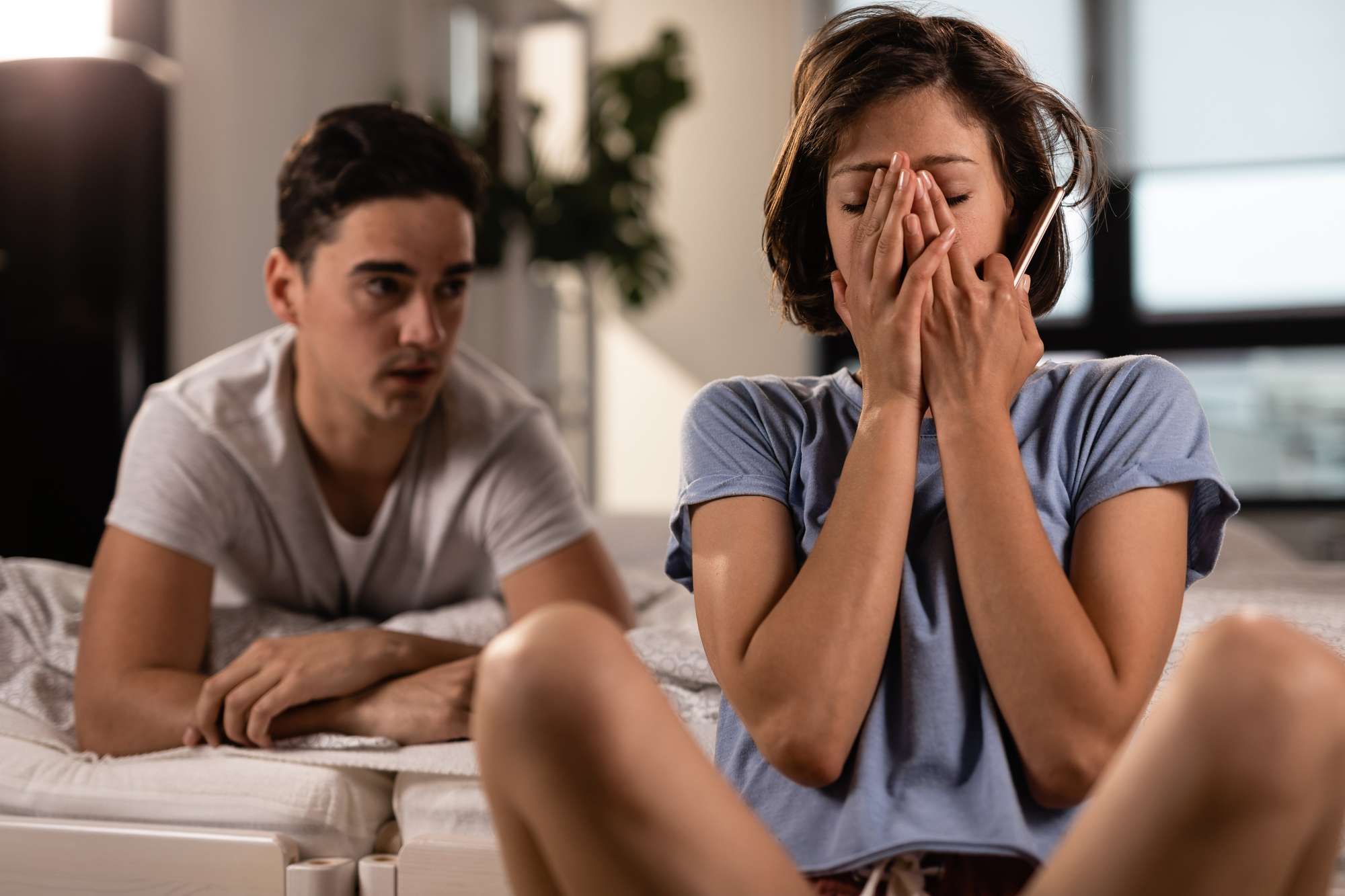 A woman sits on a bed, covering her face with her hands while holding a phone, appearing distressed. A man sitting next to her looks on with concern. The background shows a softly lit room with a plant and a window.