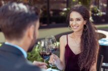 A woman with long, curly hair and wearing a burgundy dress is smiling while holding a glass of red wine. She is sitting at a table outdoors across from a man in a suit. They appear to be having a pleasant conversation.