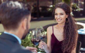 A woman with long, curly hair and wearing a burgundy dress is smiling while holding a glass of red wine. She is sitting at a table outdoors across from a man in a suit. They appear to be having a pleasant conversation.