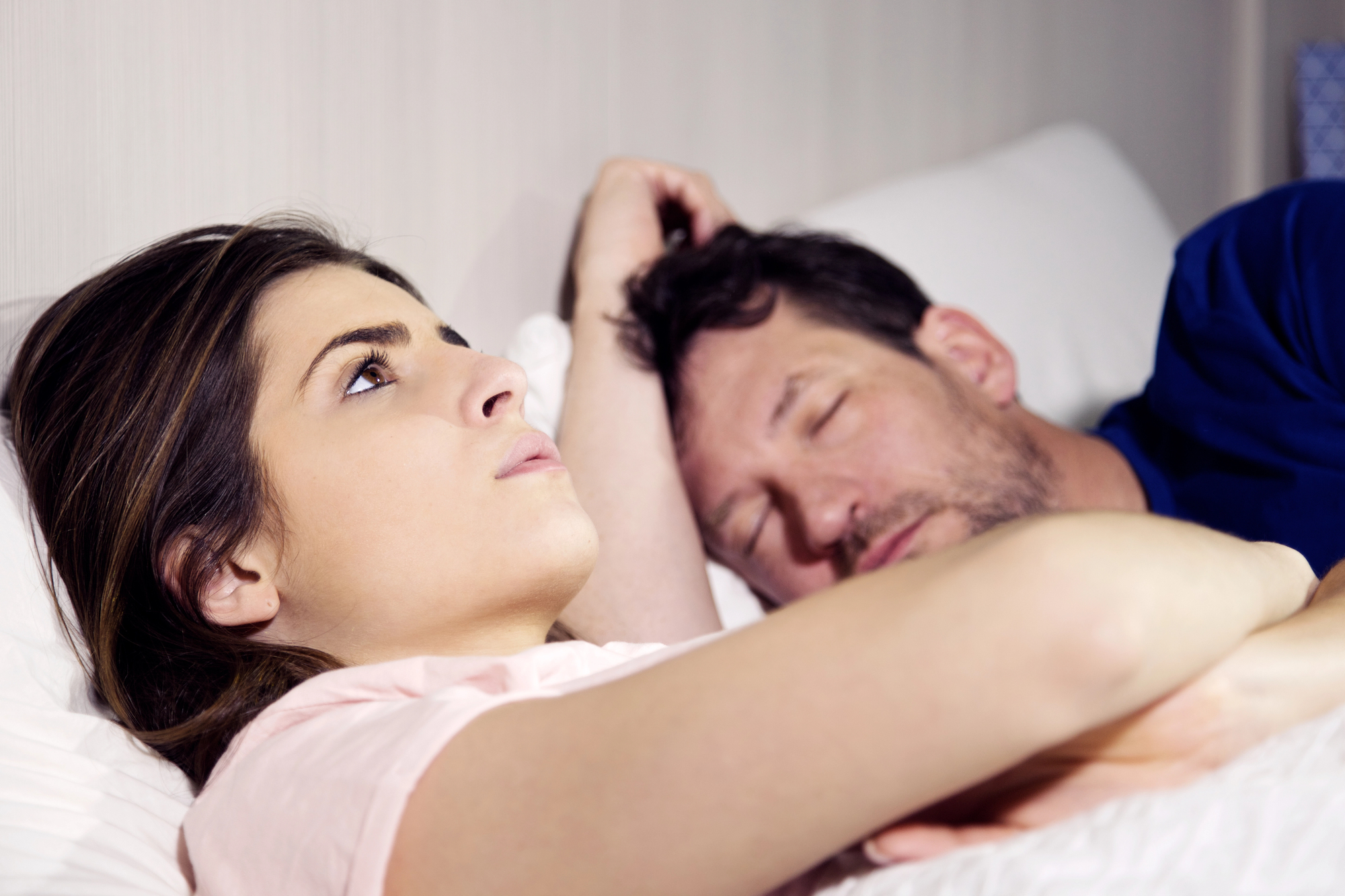 A woman lies awake in bed with a thoughtful expression, her head resting on her hand, while a man sleeps beside her with his hand resting behind his head. They are both covered by a white blanket. The woman appears pensive under the soft bedroom lighting.