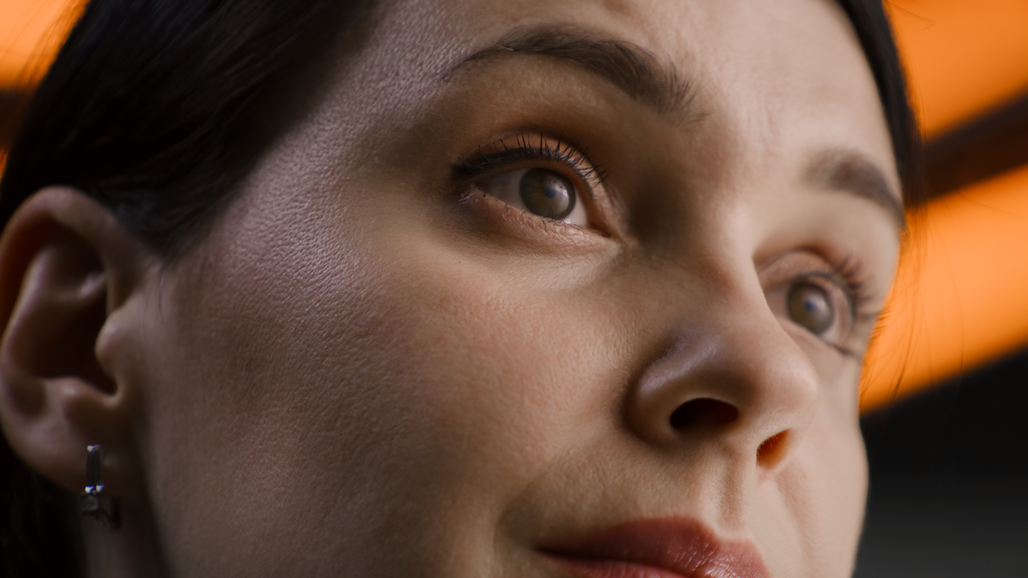 Close-up of a person staring thoughtfully into the distance. The image focuses on their eyes and part of their face, showing details of their skin texture, eyelashes, and eyebrows. The background features a warm, orange light, adding an ambient glow.