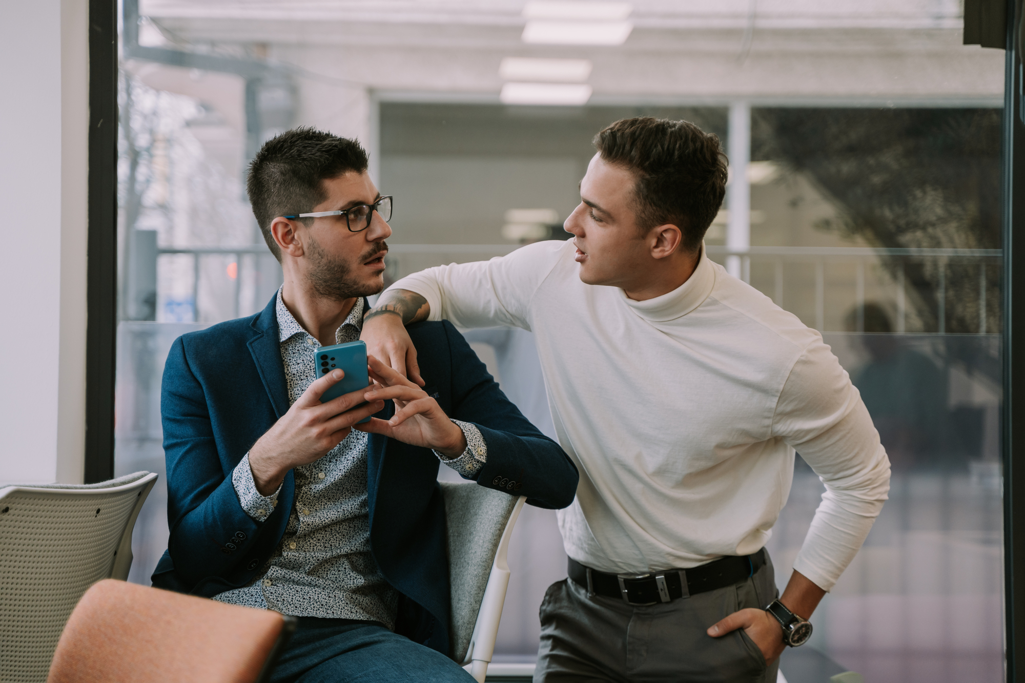 Two men having a conversation in an office. One is seated, wearing glasses, a blue blazer, and holding a smartphone. The other is standing, wearing a white turtleneck and resting his arm on the back of the chair, looking at the seated man.