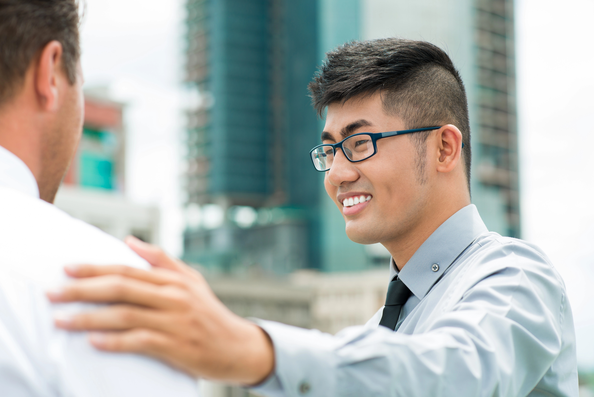 A man wearing glasses and a gray shirt with a black tie is smiling and resting his hand on another man's shoulder. They are outdoors, with modern skyscrapers in the background. The second man is only seen from the back, wearing a white shirt.