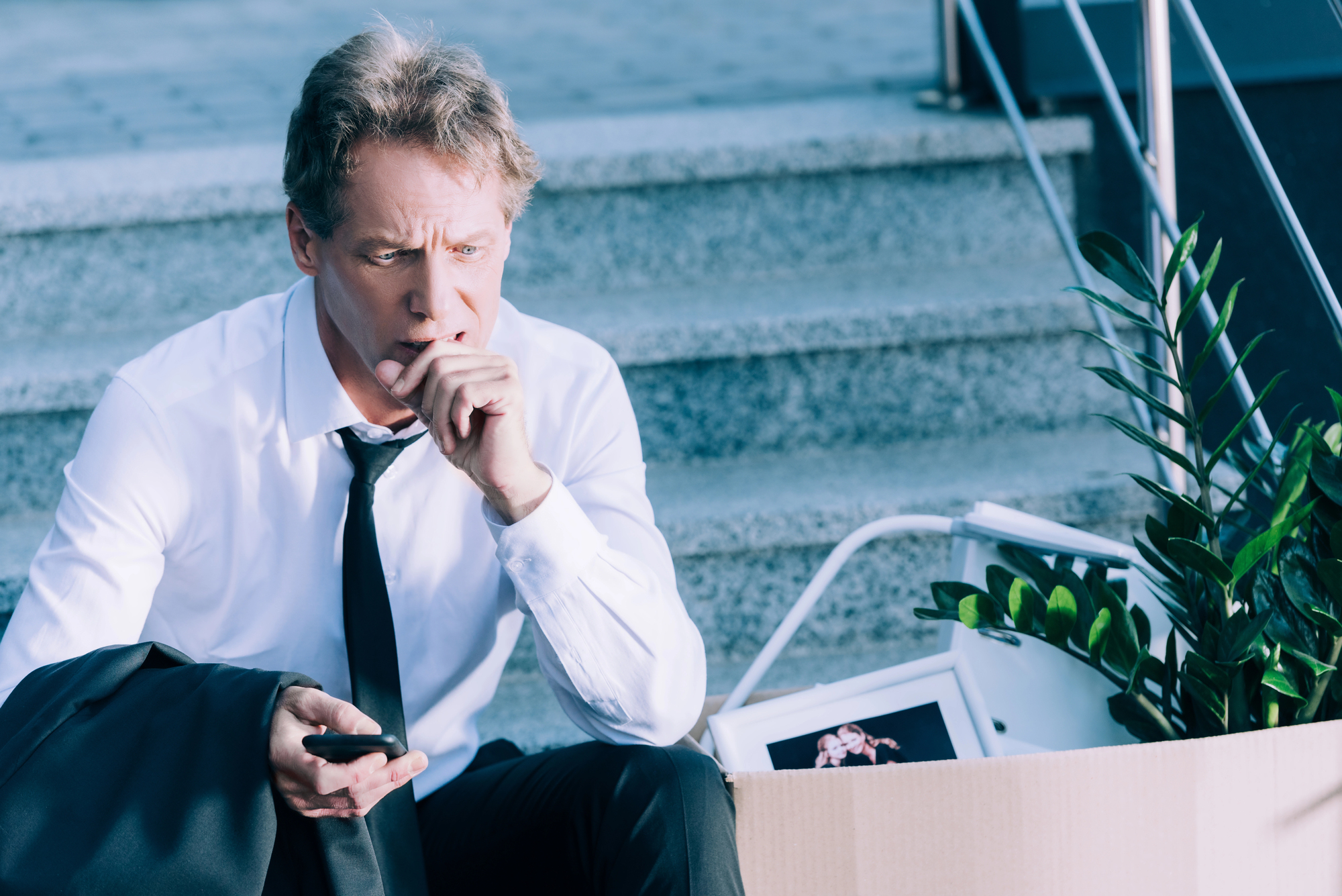 A man in a white shirt and tie sits on stairs with a pensive expression, holding his chin and a smartphone. Beside him is an open box containing a plant, a photo frame, and other personal items, suggesting he may have been laid off from his job.