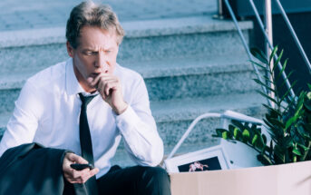 A man in a white dress shirt and black tie sits on outdoor steps, holding a smartphone in one hand and a coat in the other. He appears concerned. A cardboard box with a plant and personal items is beside him, suggesting he has recently been laid off.