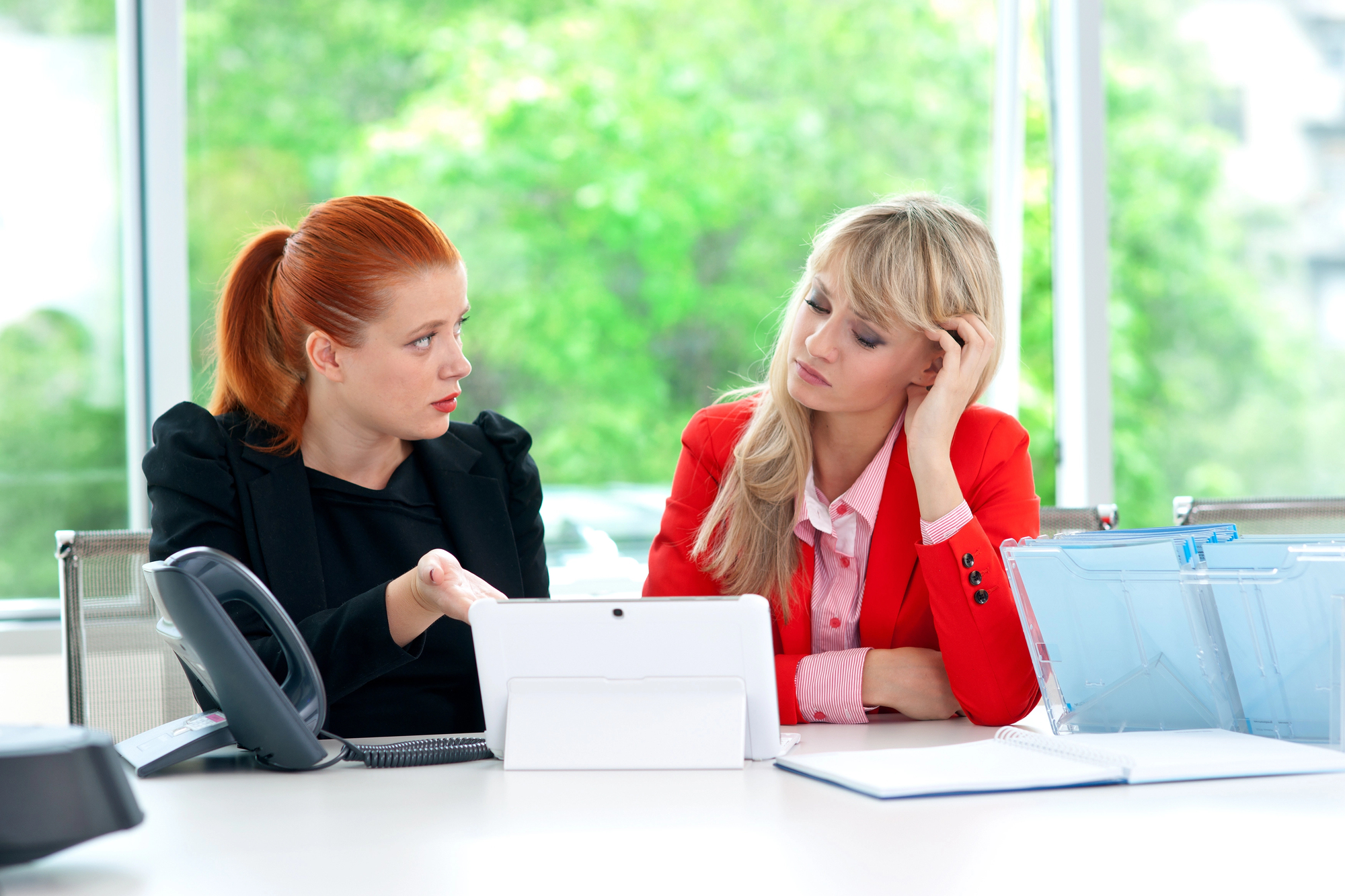 Two women are seated at a desk in an office. One, with red hair, is gesturing towards a tablet in front of them, appearing to explain something. The other, with blonde hair, looks thoughtful with her hand on her head. Office supplies and a phone are on the desk.