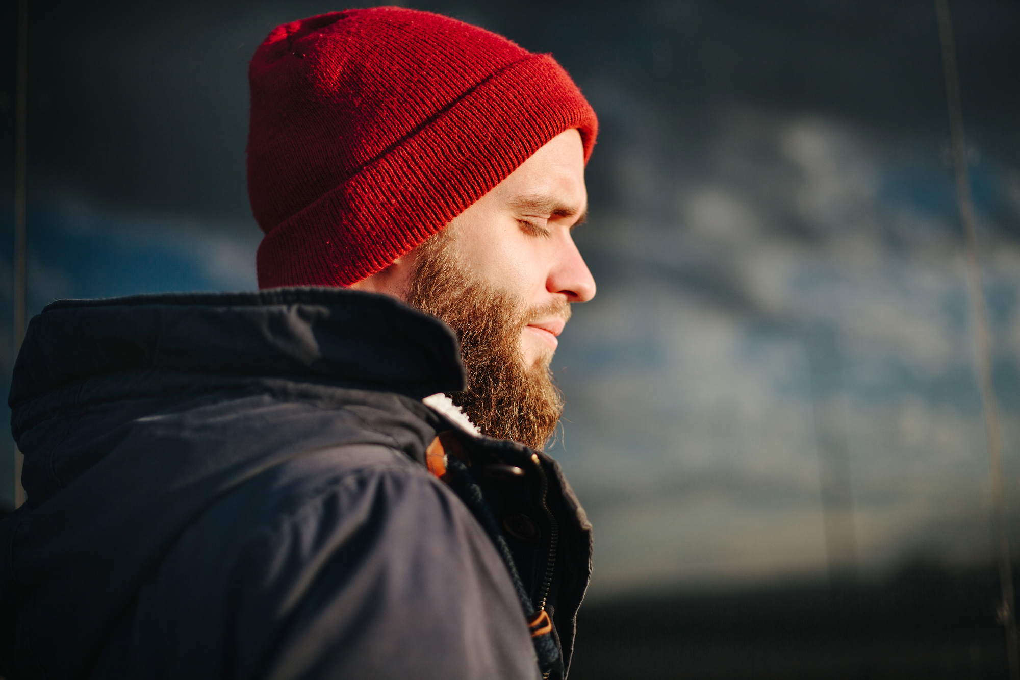 A side profile of a man with a beard wearing a red beanie and a dark jacket. He stands outdoors with his eyes closed and appears calm and peaceful. The background is blurred, showcasing a cloudy sky.