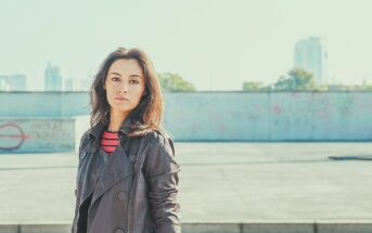 A woman with a serious expression, wearing a dark trench coat and a red striped shirt, stands in an urban setting with a defocused background featuring a graffiti-covered wall and distant buildings. The sky is clear and bright.