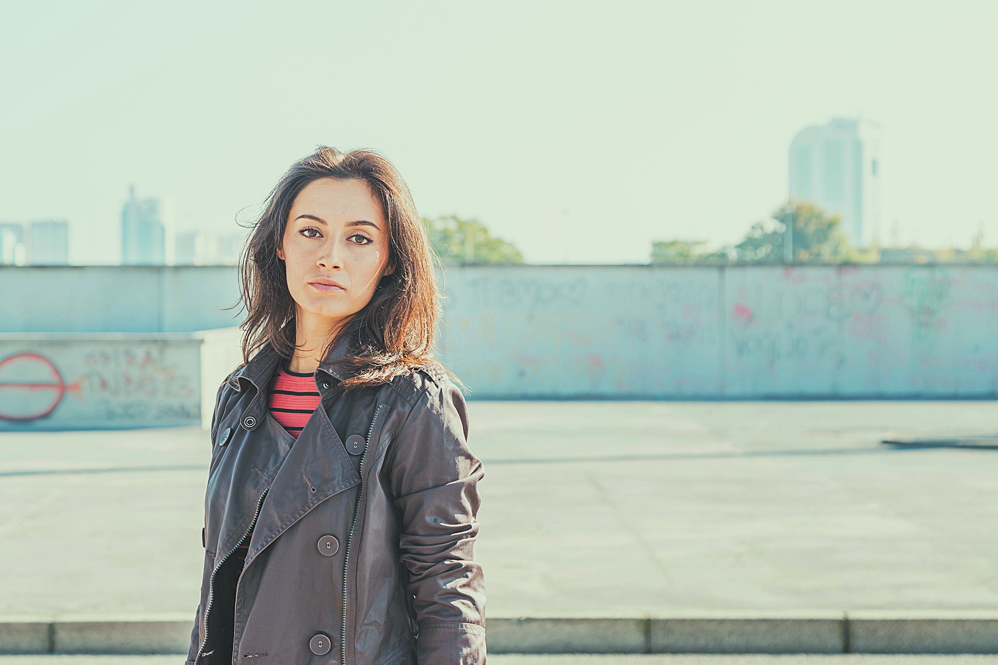 A woman with a serious expression, wearing a dark trench coat and a red striped shirt, stands in an urban setting with a defocused background featuring a graffiti-covered wall and distant buildings. The sky is clear and bright.