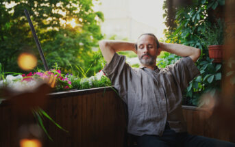 A man with a relaxed expression sits on a wooden balcony with his hands behind his head. He is wearing a loose, grey shirt. The balcony is adorned with plants, flowers, and greenery, and the background shows trees illuminated by sunlight.